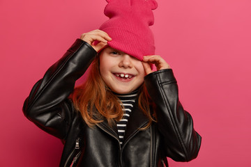 Cropped image of little girl looks from hat, hides face, wears stylish black leather jacket,...