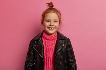 Good looking positive little kid has ginger hair combed in knot, wears rosy knitted sweater and leather black jacket, feels playful, glad after successful shopping day, stands against pink background.