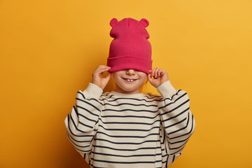 Studio shot of cheerful girl has two stick out teeth, covers half face with hat, wears casual striped jumper, has fun, gets dressed and prepares for outdoor walk, isolated on yellow background