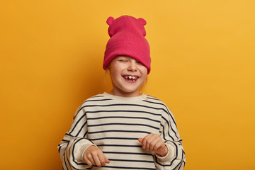 Sincere emotional child plays with new hat, dressed in striped jumper, laughs and cheers something, has funny joyful expression, playful mood, going crazy, isolated on vibrant yellow background