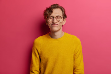 Portrait of handsome man closes eyes with pleasure, pleased to hear praising words from employer, has funny face, wears big optical glasses and yellow sweater, never stops dreaming, stands relieved