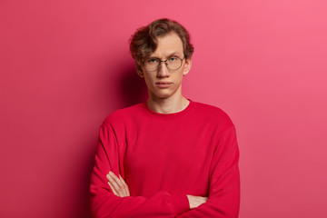 Self assured serious man keeps arms folded, looks directly at camera with confident expression, has...
