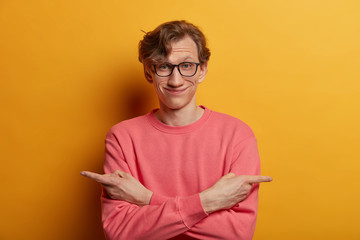 Positive European man with hesitant look, crosses arms over body, points sideways, chooses between two objects, wears spectacles and sweater. Time to make choice. Male needs advice what to select