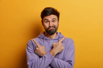 Doubtful bearded guy crosses hands and points index fingers at opposite sides, has difficulty in choosing something, purses lips with hesitation, wears hoodie, isolated on yellow background.