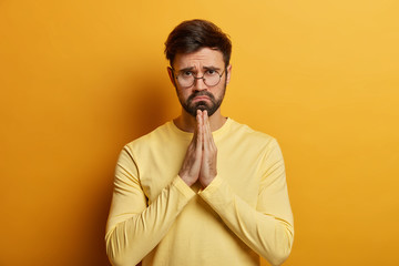 Please, forgive me. Depressed unshaven man presses palms and makes apologize, has innocent gaze, begs for help, wears spectacles, yellow jumper, looks with pity face. Male prayer asks to do favor
