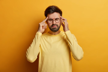 Overworked frustrated bearded man massages temples, suffers from severe migraine, closes eyes to relieve pain, wears optical glasses and casual yellow sweater, stands in studio, tries to calm down © WHstudio Leushin N