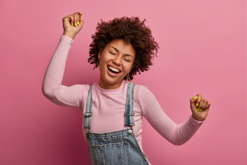 Happy dark skinned girl enjoys every moment of life, dances and moves, raises arms and clenches fists, closes eyes, has good mood, wears denim sarafan and turtleneck, isolated on pink background - Powered by Adobe