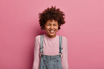 Positive young Afro American girl smiles broadly, has upbeat mood, laughs at something very funny or hilarious, closes eyes, hears crazy joke, dressed casually, stands against pastel rosy wall