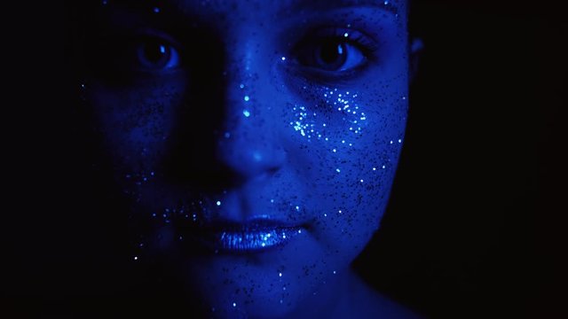 Creative glitter makeup. Aesthetic cosmetology. Woman with sparkling face opening eyes in blue glow.