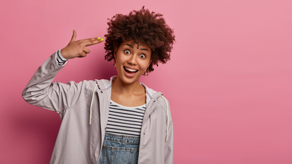 Funny glad woman with Afro hairstyle points fingers to head, going to shoot herself, wears jacket, shows suicide gesture, feels bored, tries to entertain herself, poses indoor over rosy wall © WHstudio Leushin N