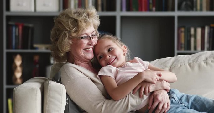Happy relaxed older granny embracing preschool granddaughter rest on couch
