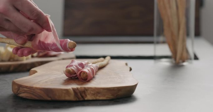 Slow motion man wrap grissini with prosciutto on olive board