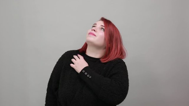 Young redhead fat lady in black sweater on grey background stylish woman poses for the camera supports face with hand