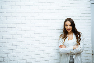 beautiful serious young business woman with dark wavy hair in white clothes crossed arms standing near white stone wall