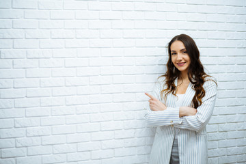 smiling business woman at work with long dark wavy hair in a white suit her arms and showing finger draws attention to white wall © ostap_davydiak