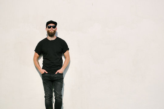 Hipster handsome male model with beard wearing black blank t-shirt and a baseball cap with space for your logo or design in casual urban style