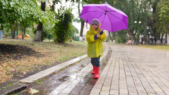 4k slow motion video of cute little boy in raincoat and rubber boots holding umbrella and walking in auutmn park while raining