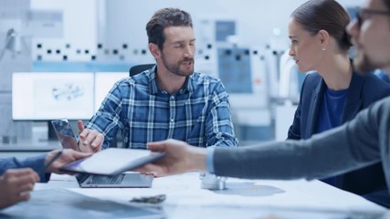 Modern Factory Office Meeting Room: Multi-Ethnic and Diverse Team of Engineers, Managers and Investors Talking Sitting at Conference Table, Analyzing Blueprints, Showing Information on Laptop Computer