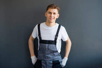 Portrait of a young, smiling, professional worker in gray overalls and white T-shirt, standing on a gray background.