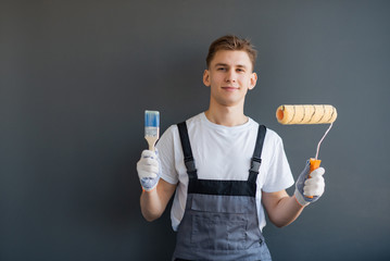 Handsome young smiling worker with paint roller and brush on gray background