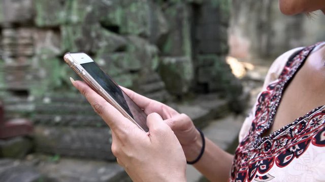4K, Close up of a tourist asian woman holding and typing on mobile phone in famous landmark of Ta Prohm ancient Hindu temple. Girl check the smartphone and surfing in internet in Cambodia vacation-Dan
