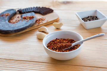 Spicy and hot Crushed dried chili peppers in white bowl - Tucana ljuta paprika in front of special ironed sausage, garlick and a bowl of mixed pepper on a wooden table