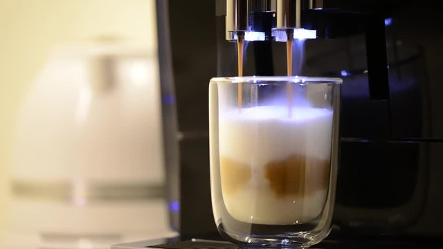 Close-up of the process of making latte coffee from modern coffee machine, poured in clear glass cup. Coffee machine is making fresh hot latte macchiato with milk foam.