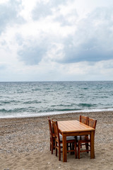 Fototapeta na wymiar A wooden table with chairs on a pebble sandy beach bay with view to wavy sea and the horizon on a cloudy day. 