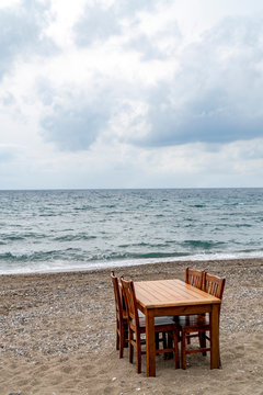 A wooden table with chairs on a pebble sandy beach bay with view to wavy sea and the horizon on a cloudy day. 