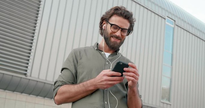 Casually dressed Young Attractive man enjoying Music on his Smartphone. Downloading popular Songs at Mobile App. Using modern white Headphones. Having eyeglasses and Beard.