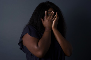 Anxiety - a conceptual image of a beautiful young African  woman covering her face with her hands and peering out with one eye between her fingers standing indoors. Scared from something or someone.