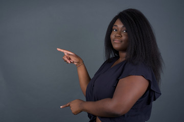 Positive attractive young African American female, points aside with cheerful and surprised expression with mouth opened, shows something amazing standing indoors. Advertisement concept.