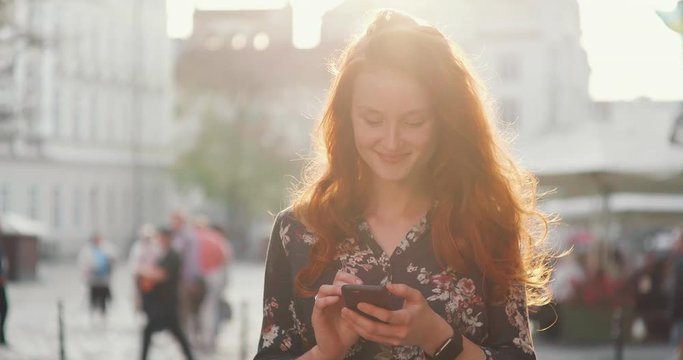 Young Girl with Red Hair dressed in black Shirt with flower print Walks down the crowded street. She is reading something in her phone and Smiling. Sun shining very brightly this day.