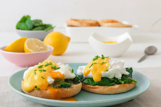 Eggs Florentine, English muffins, grilled ham, poached eggs, Hollandaise sauce, chive herbs, lemon, cup of coffee. White wooden table with breakfast.
