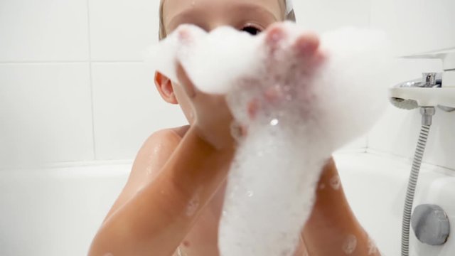 Closeup slow motion video of happy smiling boy washing in bath and blowing soap foam in camera