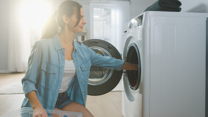 Beautiful Smiling Brunette Young Woman Sits in Front of a Washing Machine in Homely Jeans Clothes. She Loads the Washer with Dirty Laundry. Bright and Spacious Living Room with Modern Interior.