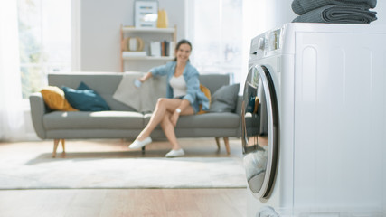 Shot of a Modern Technologically Advanced Washing Machine Working in a Bright and Spacious Living Room with Cozy Interior. Young Woman in Home Clothes is Using a Smartphone in the Background.