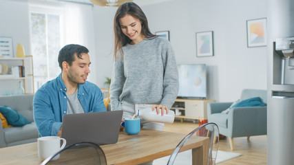 Beautiful Young Couple Talking in a Kitchen at Home. Man is Working on a Laptop, Girl d Pours Dairy Milk Yoghurt into the Mug. Thay are Happy and Laugh. Room has Modern Interior.