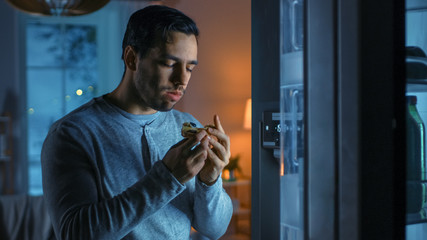 At Night in the Kitchen Handsome Attractive Young Man is Eating a Leftover Pizza from the Fridge. He is Hungry and Feels Satisfied.