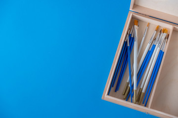 Flay lay, top view table desk. Set of different artist brushes in a wooden box on a blue...
