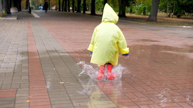 Slow motion video of little boy in red rubber boots and yellow raincoat jumping in big puddle after rain