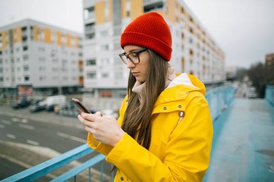 Girl in yellow raincoat checking up social media and news online. Millennial girl using mobile phone outdoors