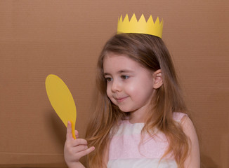 Portrait of emotional little girl with crown a yellow paper holding a mirror. handmade. cardboard background