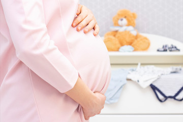 pregnant woman in a dress holds her hands on her stomach on the background of things for the newborn and a toy bear. Pregnancy, motherhood, preparation and expectation concept. Close-up indoors.