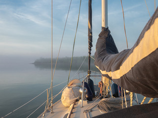 Bow of a sailboat on a calm, foggy day with sails covered and gear on deck facing a hazy forested...