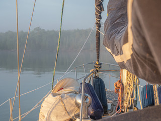 Looking forward to the bow of a sailboat in still water with sails down and gear on deck facing a hazy forested shoreline obscured by fog.