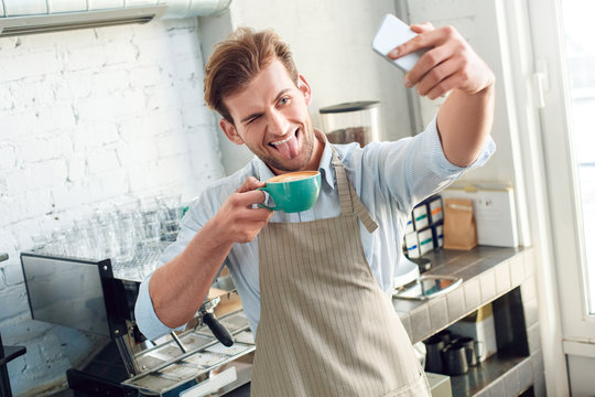 Coffee Passion. Young barista at coffee shop with cup of hot coffee taking selfie on smartphone showing tongue playful - Powered by Adobe