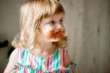 Cute curly redhead baby child shows the hands smeared by chocolate.Cute girl eats a bell at home. all dirty, stained with sweets. July 7 is chocolate day.Indoor plants, green leaves.