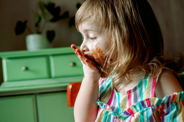 Cute curly redhead baby child shows and licks hands smeared by chocolate.Cute girl eats a bell at home. all dirty, stained with sweets. July 7 chocolate day.Indoor plants, green leaves.