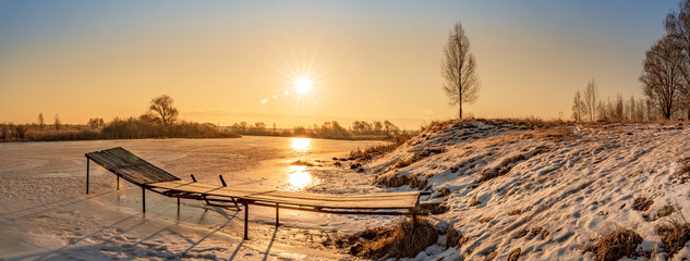Wide angle rural panorama of colorful winter sunrise over the frozen river, birch tree and wooden pier with azure sky. . Travel destination Russia